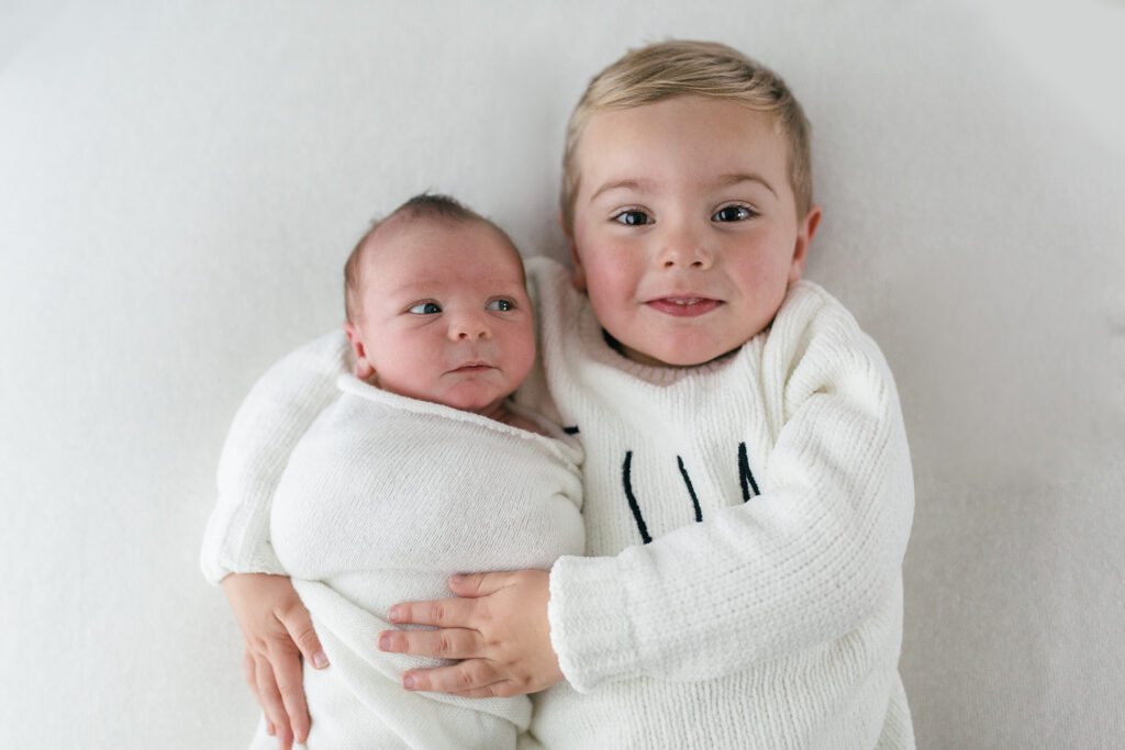 newborn baby poses with his older brother and looks at him whilst his older brother is smiling 