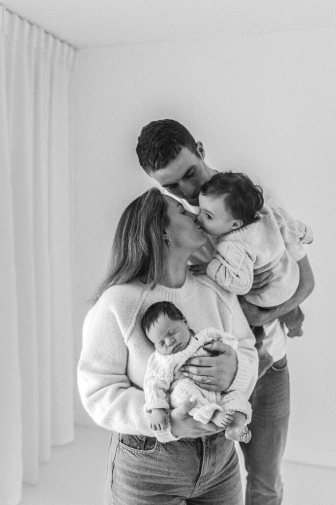 a family of four pose together for a photograph, toddler leans over ans kisses mum on the lips as dad holds him