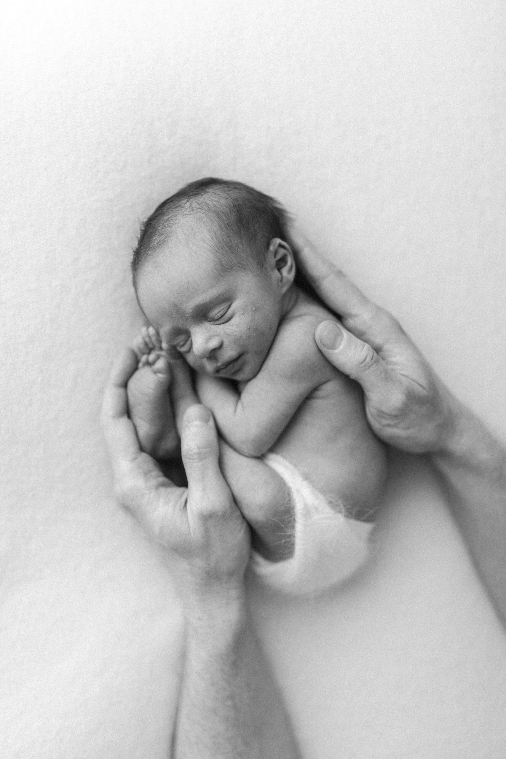 Black and white photograph of newborn baby asleep on a beanbag, dads hands holding him