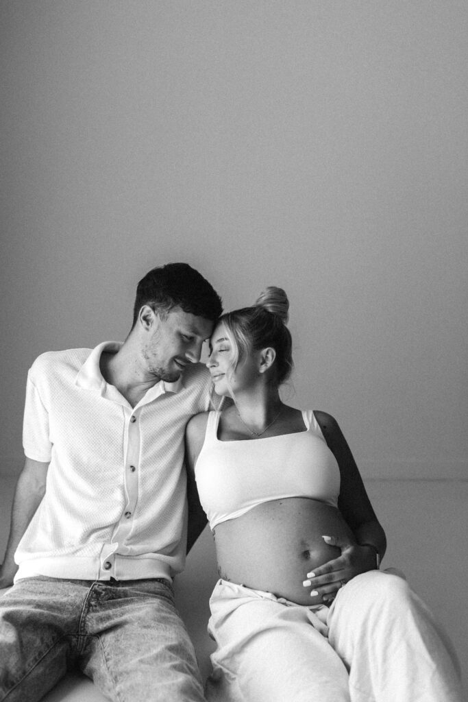 Pregnant woman and her husband are sitting on the floor, leaning in together and smiling 