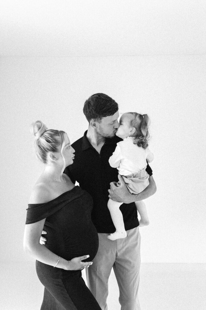 Black and white image of a family of three, mum holder her bump whilst the toddler leans over and kisses dad 