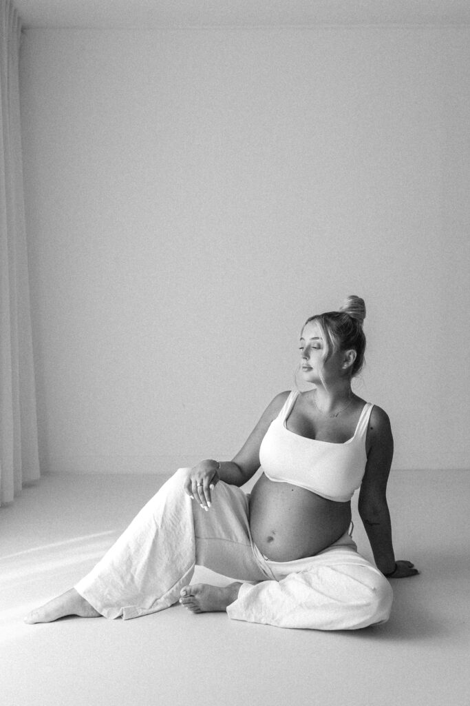 Pregnant women sits on the floor of a white photography studio, wearing white wide legged trousers and white top. The image is edited in black and white and is a very editorial style 