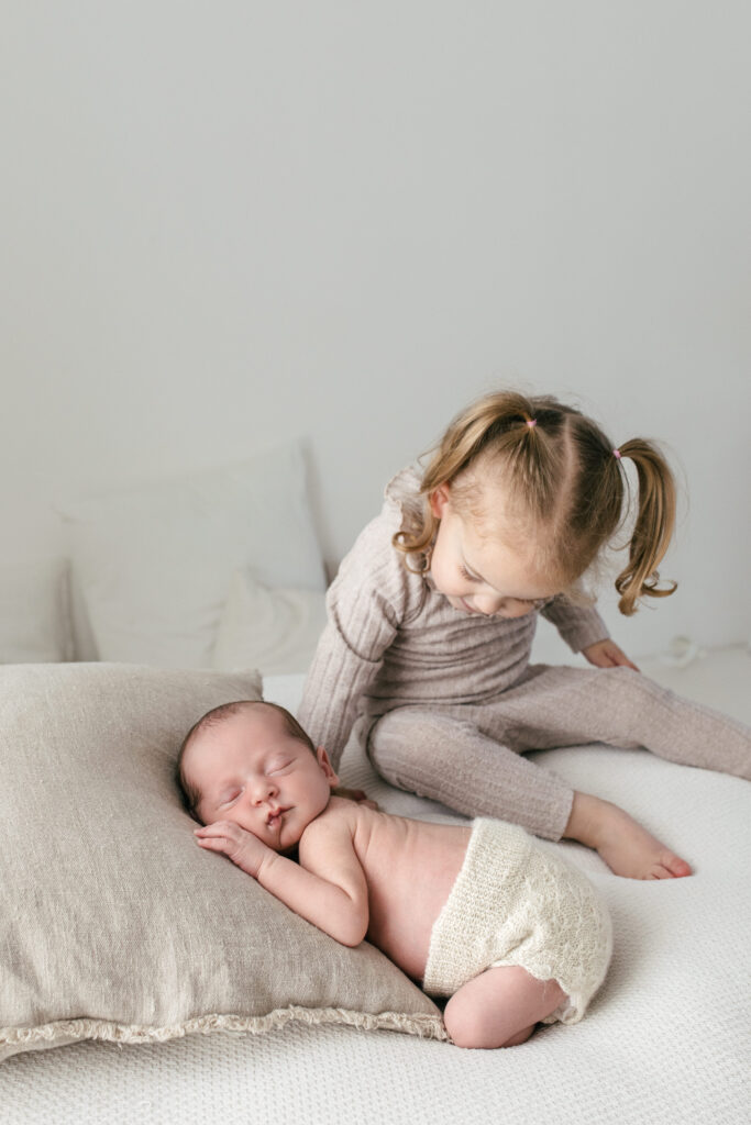 Newborn baby boy sleeps next to his sister, sister looks down at him and smiles