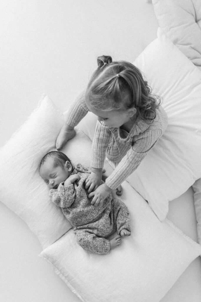 Toddler and baby sit together, toddler is tickling babies tummy 