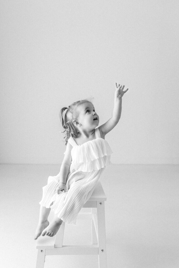 Little girl sits on a stool and reaches up to the ceiling 