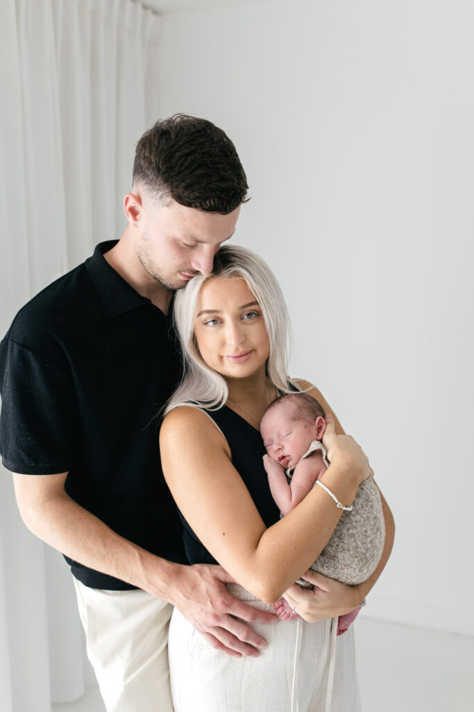 Dad, mum and newborn baby have a cuddle, dad is looking down to mum and mum is looking at the camera 