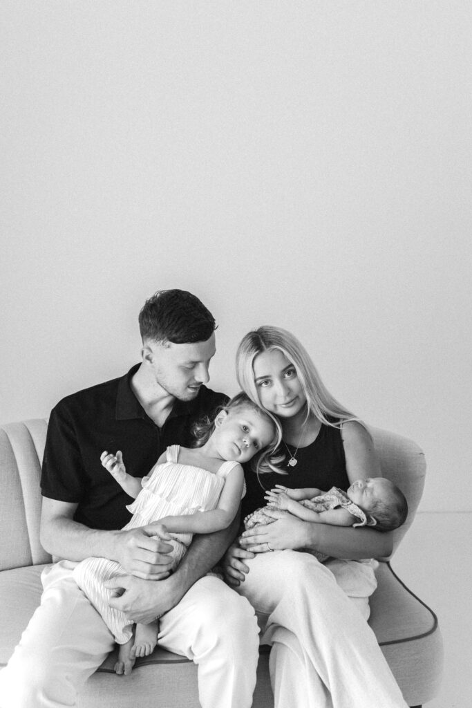 Family of four, mum and dad and toddler and newborn baby sit on a sofa together. The toddler and mum have thier heads leaning together and are looking at the camera 