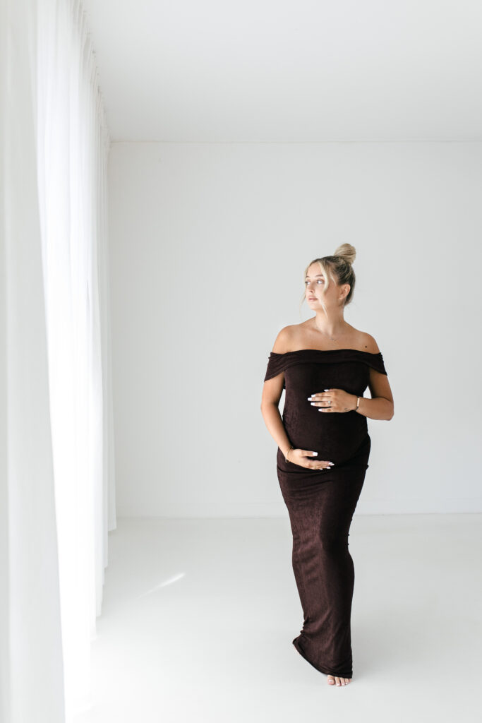 Pregnant woman, wearing a brown floor length gown poses for a photograph in a white studio, whilst holding her pregnancy bump