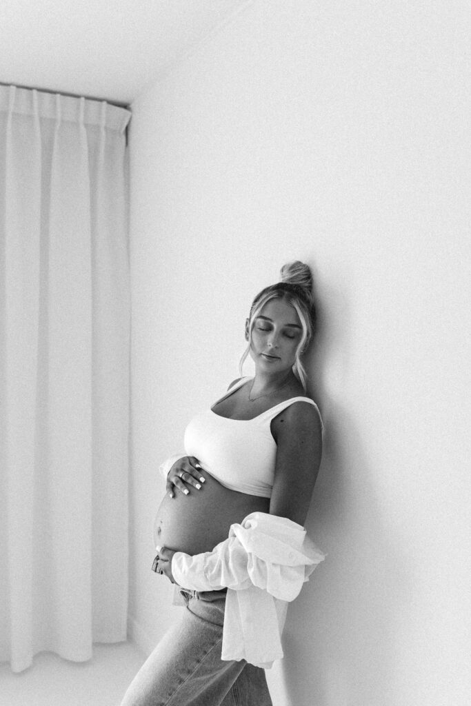 Pregnant mother leans against the wall in a white studio, holding her bump. She is wearing jeans and a white, open shirt 