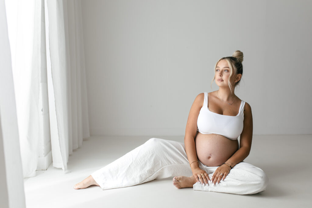 Pregnant woman poses fro photograph wilst sitting on the floor in an all white studio