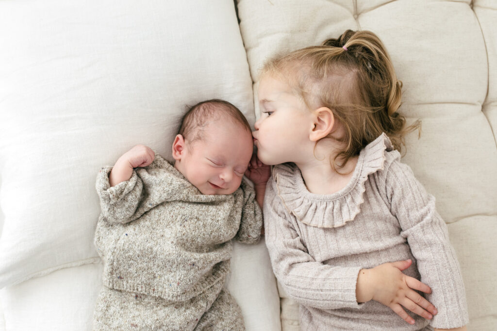 Toddler sister kisses newborn baby on the forehead as he smiles 