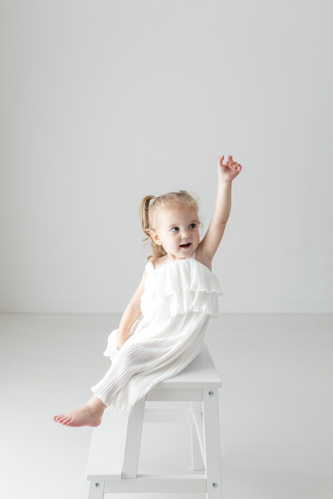 Little girl wearing all white, sitting on a stool and lifting her arms up 